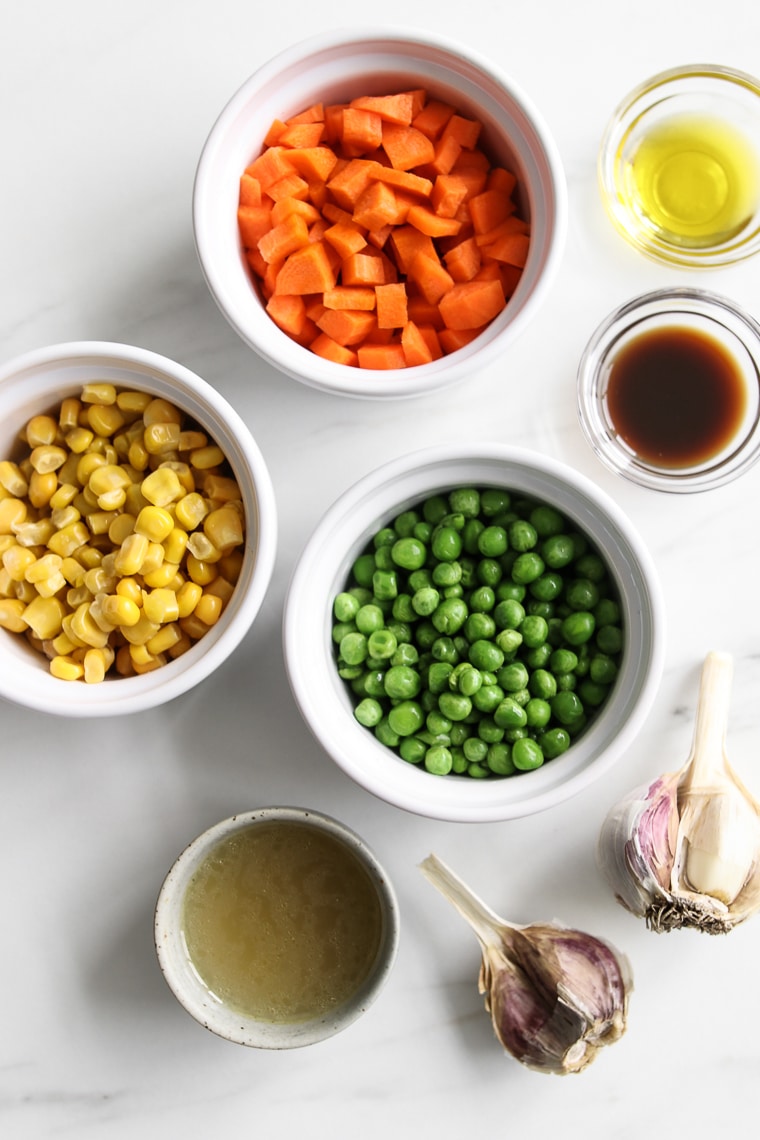 Some ingredients for traditional cottage pie: Small bowls of diced carrots, corn kernels, green peas, worcestershire, beef stock and cloves of garlic 