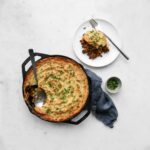Beef Shepherd's Pie in a cast iron skillet next to a plate with a piece of shepherd's pie and small bowl of parsley