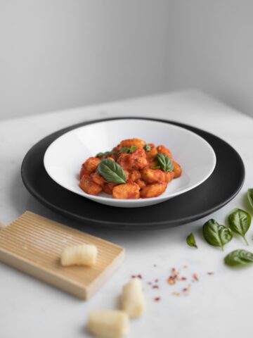 Gluten-free gnocchi with tomato sauce in a white bowl, on top of a black plate. Uncooked gnocchi and basil are placed around the plate. Get the recipe at chefsouschef.com