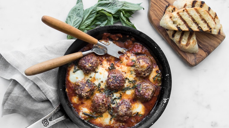 Close up image of spicy meatballs in a skillet with melted cheese, serving utensils, fresh basil, and two slices of grilled bread