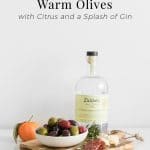 Tall image of warm olives in a bowl on a wooden board with parmesan cheese, salami, rosemary and a bottle of gin