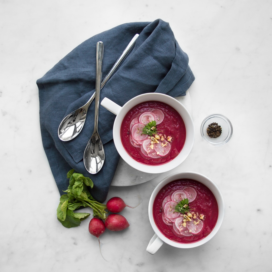 Close up of two mugs of beet soup with radishes and a blue napkin with two spoons.