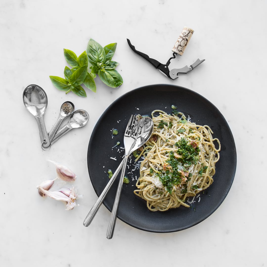 Overhead image of plated Pesto Carbonara on a black plate with a fork and spoon, laying on marble with basil, garlic cloves, and a corkscrew with a cork