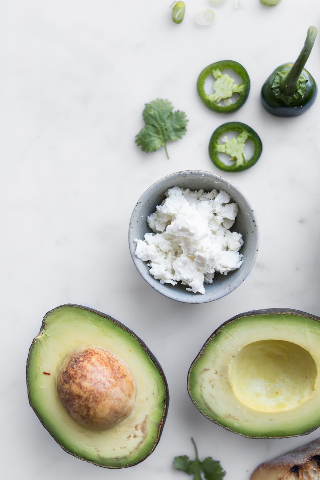 Halved avocado, bowl of crumbled cheese, cilantro leaf, and jalapeno slices on a table