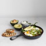 Side view of Mexican Shakshouka in skillet next to sliced bread and avocado