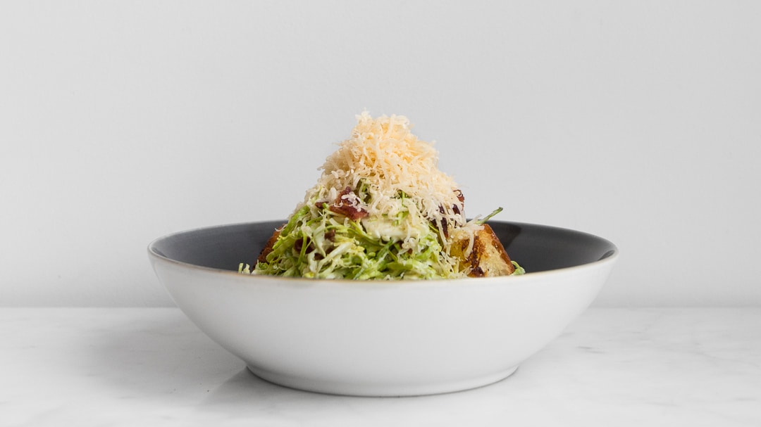 A bowl with filled with brussels sprout caesar salad with grated gouda on top