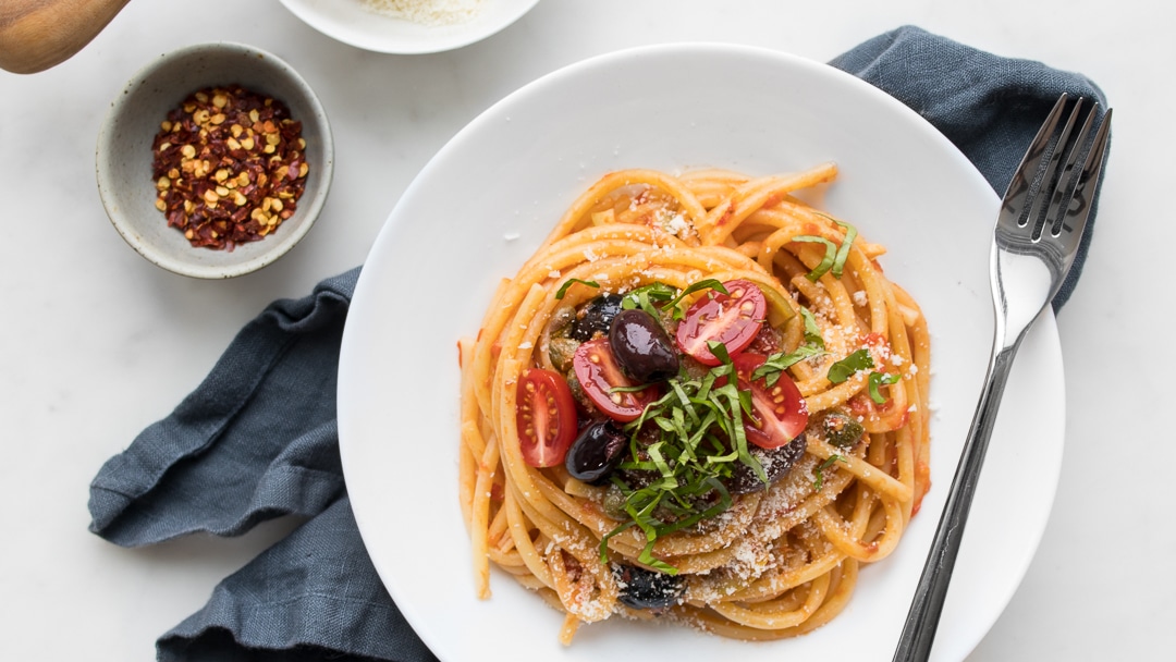 Spaghetti Puttanesca on a white plate with small bowl of chilli flakes