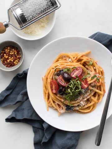 Spaghetti Puttanesca in a bowl next to chilis and parmesan