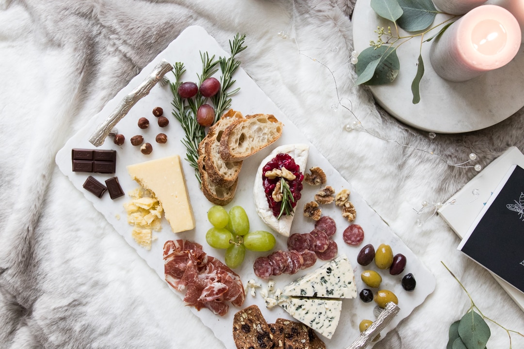 Comforting cheeseboard styled on a marble tray with cheese, meat, chocolate, olives, fruit, and crackers.