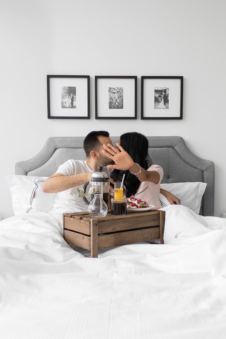 Mystique's hand blocking a view of Philip and Mystique kissing, while laying in their bed with a wooden crate with coffee and french toast. The bed has a grey headboard and 3 black and white pictures hanging above.
