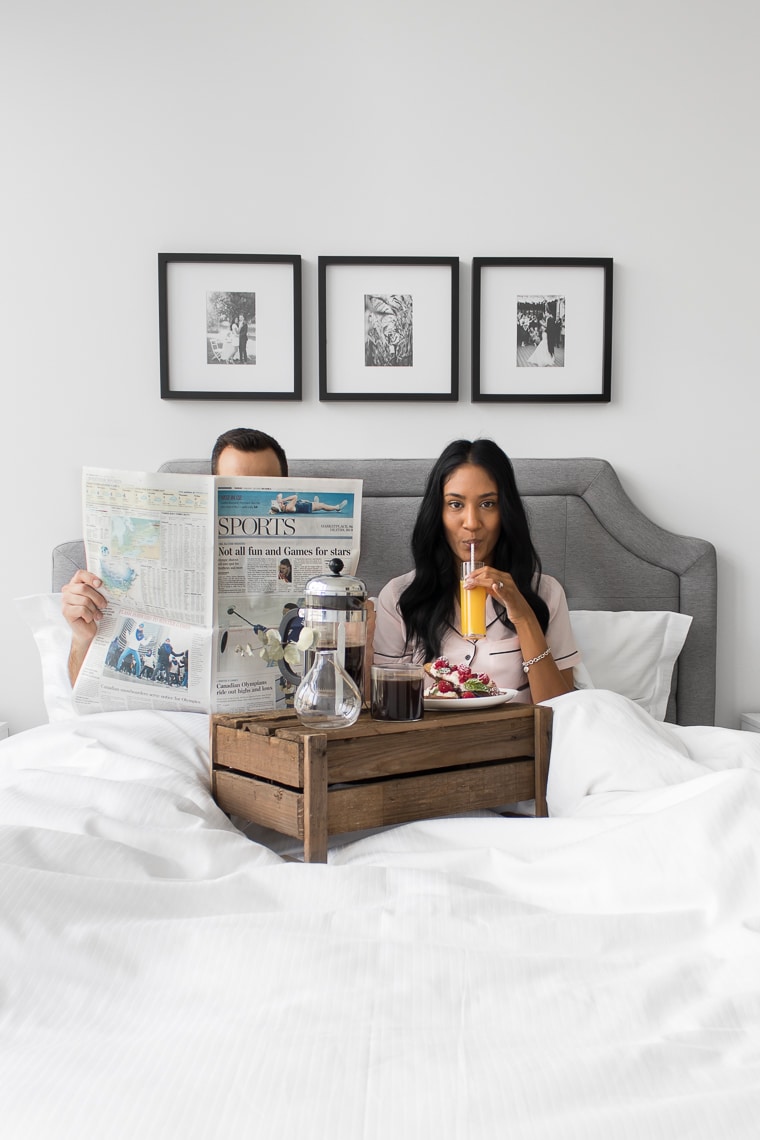 Philip reading a newspaper with his head poking above and Mystique sipping on orange juice looking at the camera while sitting up in bed behind a wooden crate with french toast and coffee.