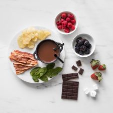 Overhead of Dark Chocolate Fondue for Two in a small cast iron pot surrounded by a chocolate bar, mint leaves, chips, strawberries, and bowls of raspberries and blackberries