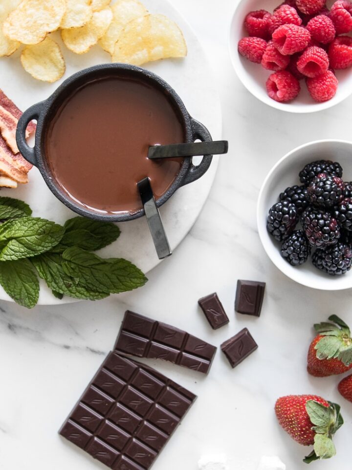 Dark Chocolate Fondue for Two in a small cast iron pot surrounded by a chocolate bar, mint leaves, chips, strawberries, and bowls of raspberries and blackberries