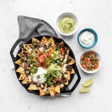 Loaded nachos in a cast iron pan with bowls of guacamole, salsa, and sour cream