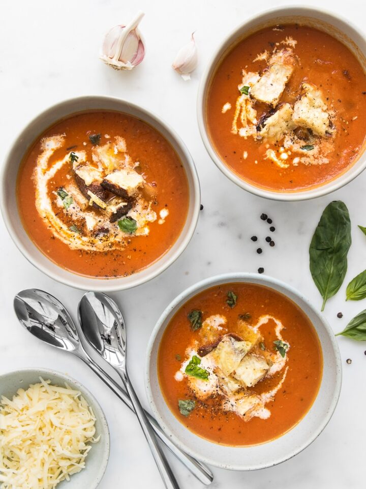 Close up of three bowls of Roasted Tomato Soup with Grilled Cheese Croutons next to a small bowl of grated cheese and spoons