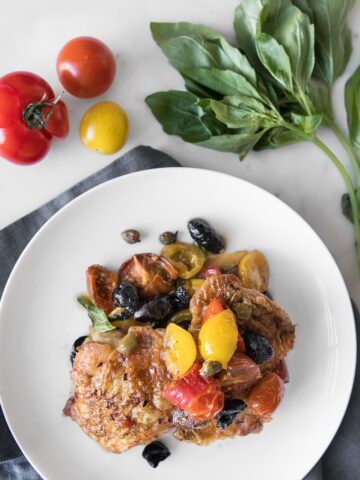 Plated Chicken Puttanesca on a white plate next to fresh basil and tomatoes