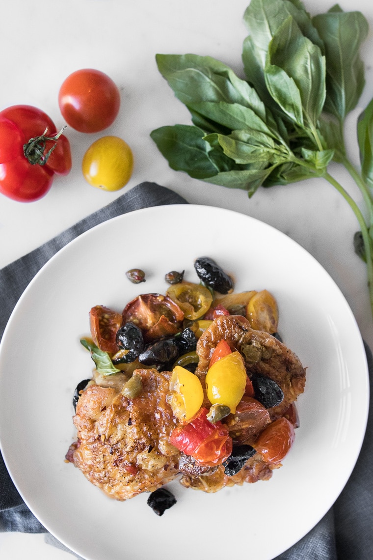 Plated Chicken Puttanesca on a white plate next to fresh basil and tomatoes