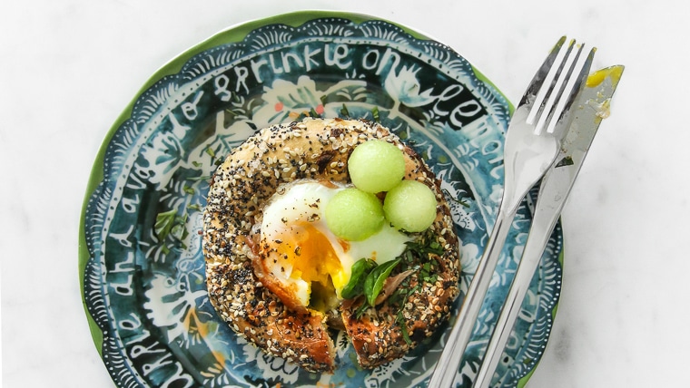 Egg in a hole bagel with prosciutto and melon on a green plate with a fork and knife