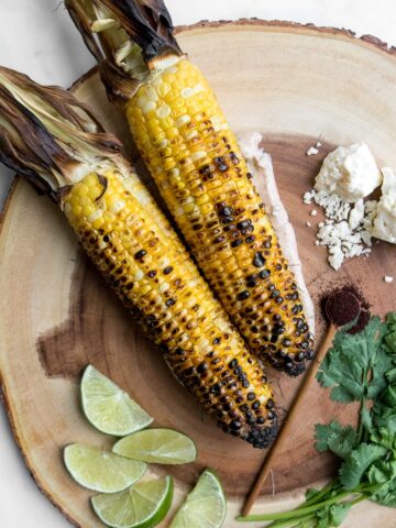 Two grilled corn cobs on a wooden board with lime wedges, cheese, and cilantro