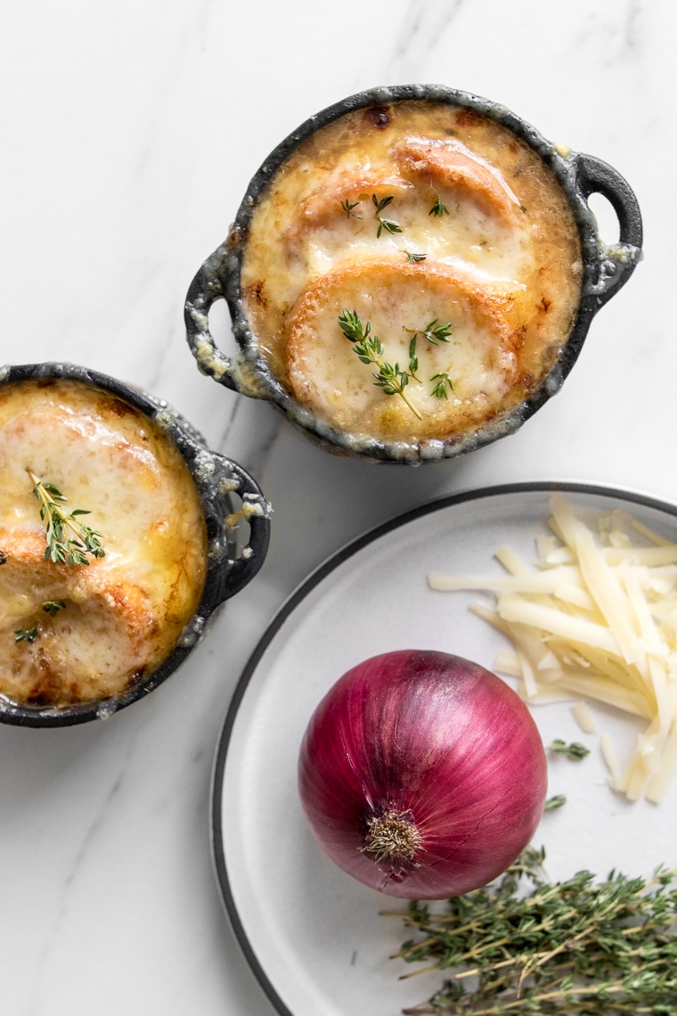 Two bowls of the French Onion Soup next to a plate with onions, cheese and thyme