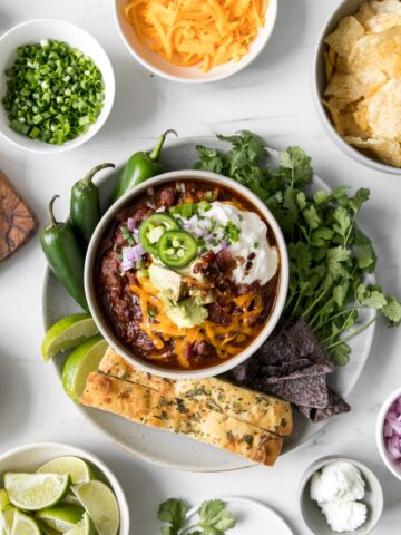 Homemade chili in a bowl topped with cheese, sour cream, avocado, and jalapeno peppers surrounded by bowl of various chili toppings