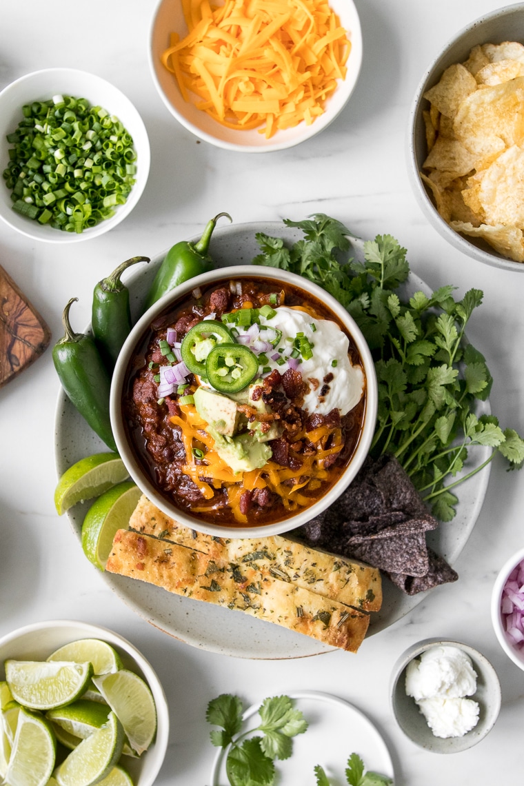 Homemade chili in a bowl topped with cheese, sour cream, avocado, and jalapeno peppers surrounded by bowl of various chili toppings