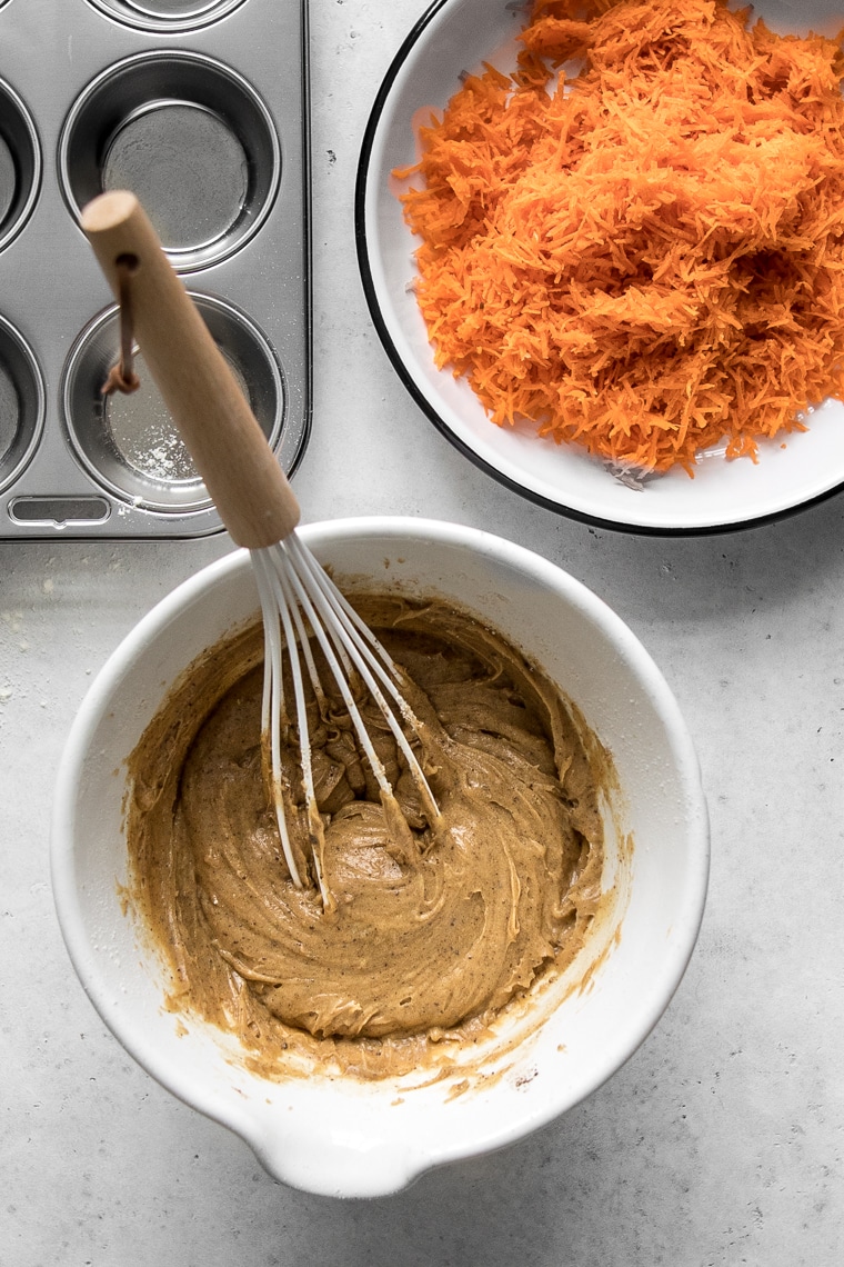 Muffin batter in a bowl, next to grated carrots and a muffin pan