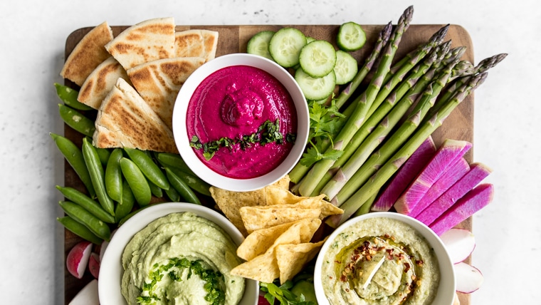 Close up of large cutting board with three dips, vegetables and pita bread