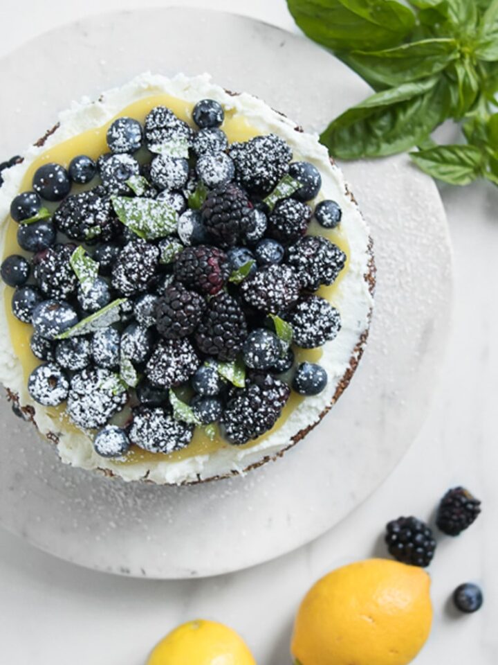 Cake with fresh berries on top next to lemons, blackberries, and basil