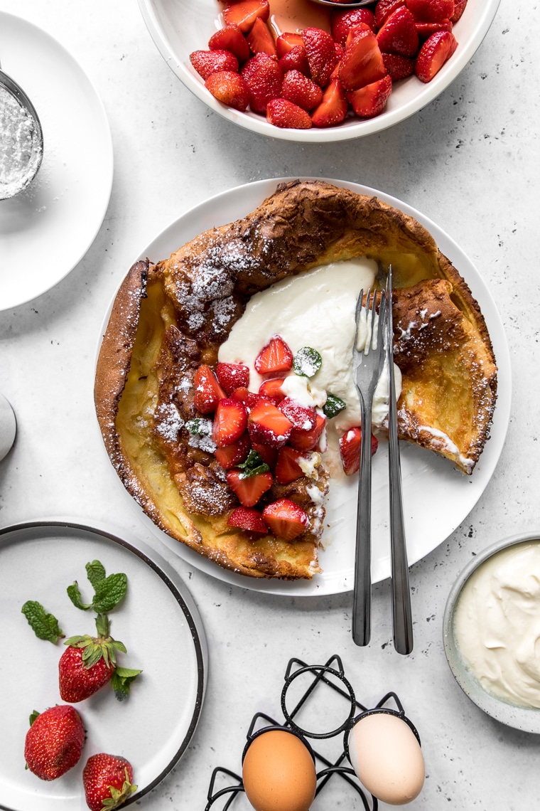 Dutch Baby with Whipped Sour Cream and Strawberries with slice taken and surrounded by strawberries and eggs