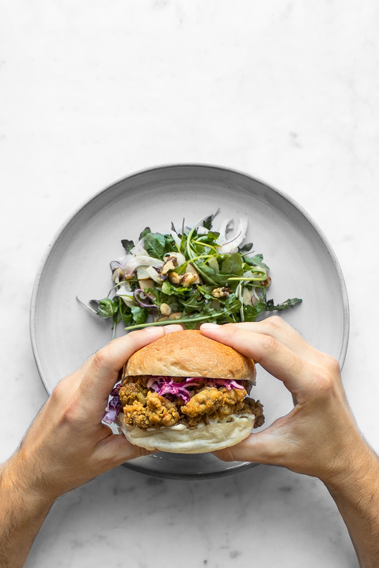 Hands holding a fried chicken sandwich above a plate with salad