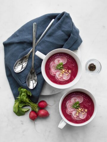 Two bowls of beet soup styled with blue napkins, spoons and a bunch of radishes