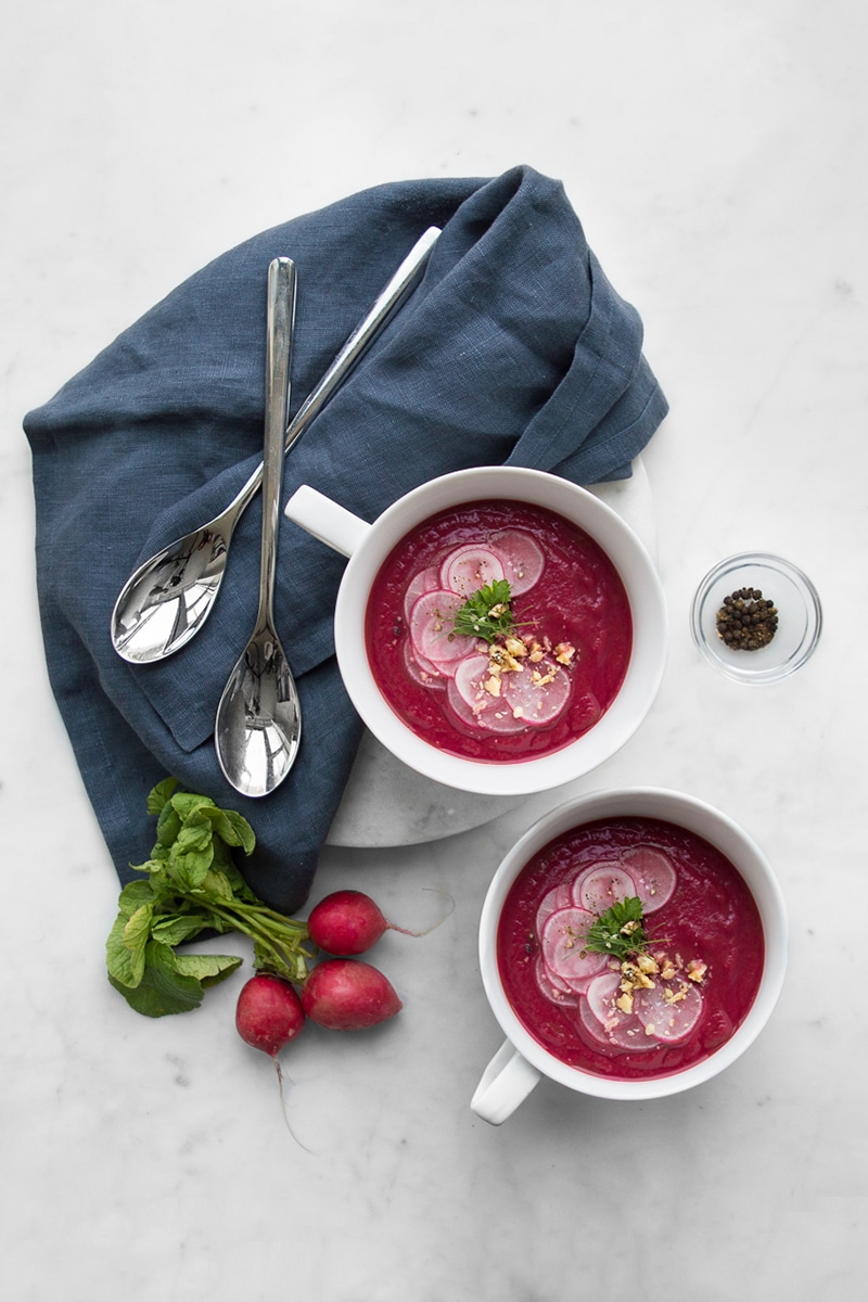 Two bowls of beet soup styled with blue napkins, spoons and a bunch of radishes