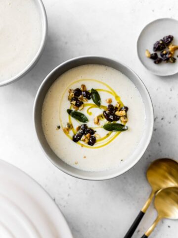 Bowl of Creamy Cauliflower Soup with fried sage, walnuts and raisins on top with gold spoons and pot lid