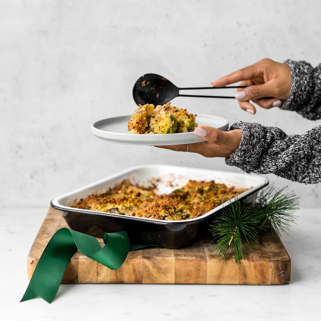 Hands shown serving brussels sprout gratin from roasting pan set on wooden board