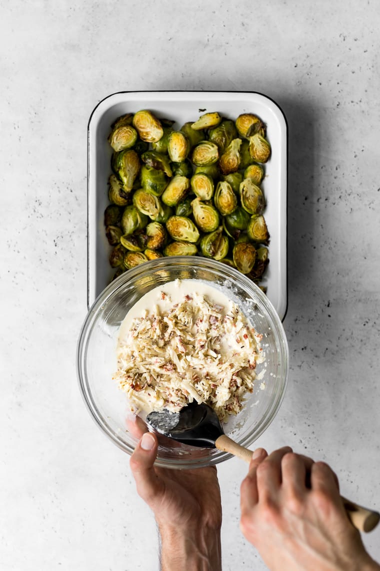 bowl with gratin mixture being held above baking pan of roasted brussels sprouts