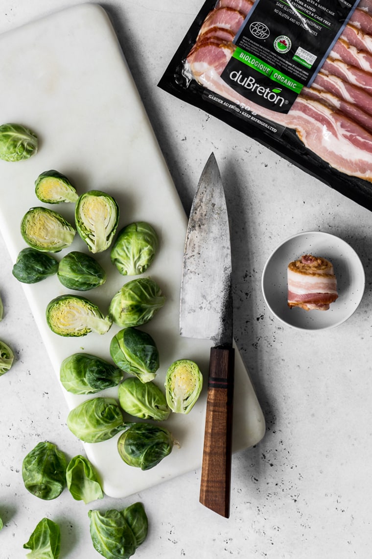 Marble board with sliced brussels sprouts  with knife and next to package of bacon and rolled piece of bacon in a small bowl