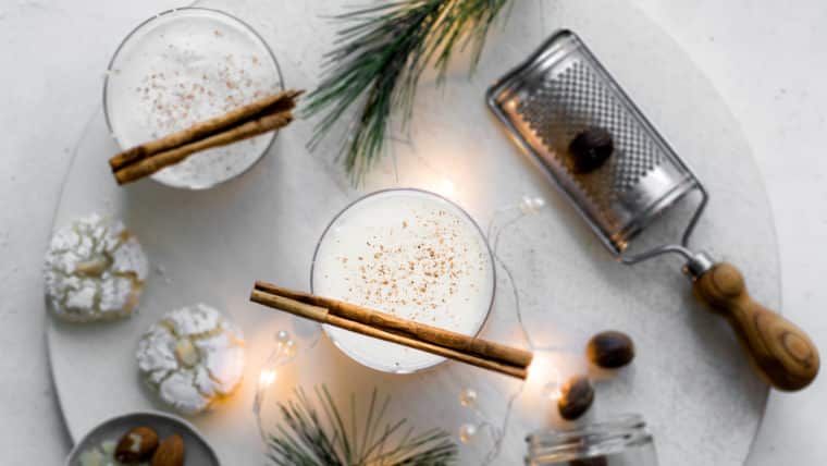 Two glasses of eggnog with cinnamon sticks, shot from above with nutmeg grater, cookies and pine leaves