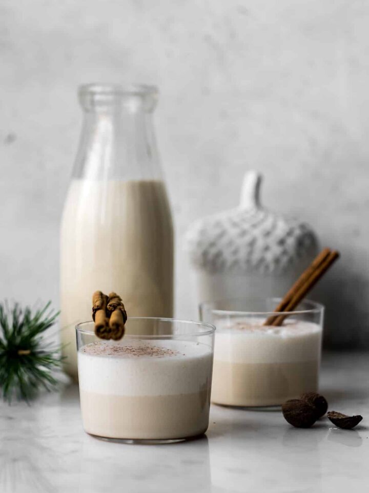 Eggnog in small glasses with cinnamon sticks, jug of eggnog and holiday decor