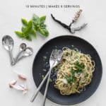 Black plate with walnut pesto pasta, basil, measuring spoons, garlic and the recipe title