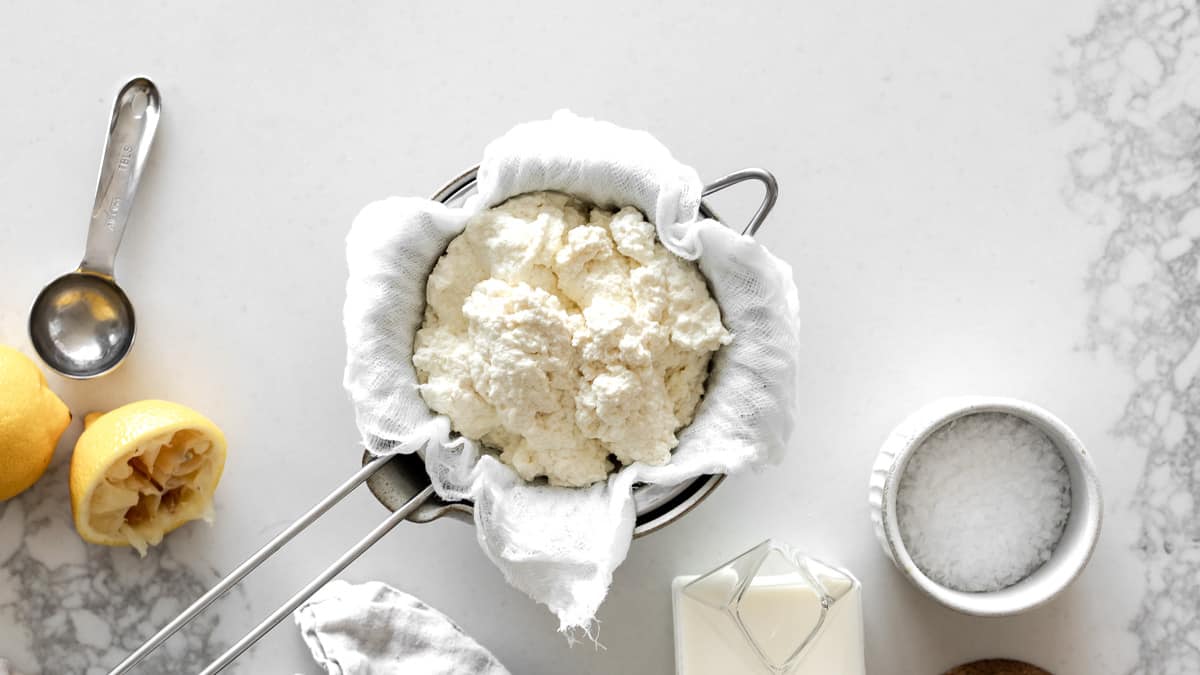 Homemade Ricotta Cheese in cheesecloth with lemons, measuring spoon, salt and milk around it.