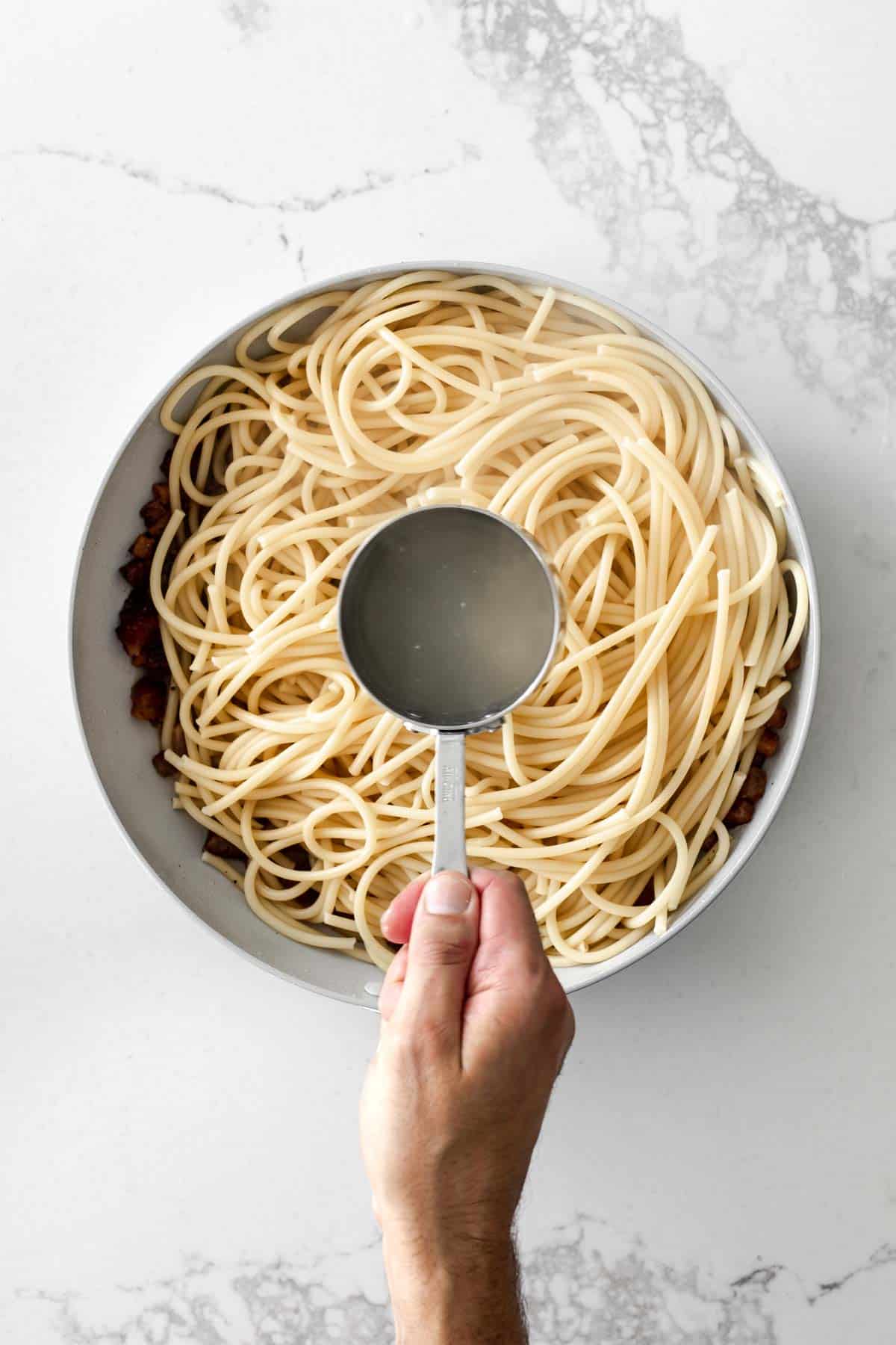 Hand holding a measuring cup with starchy pasta water above a skillet of spaghetti