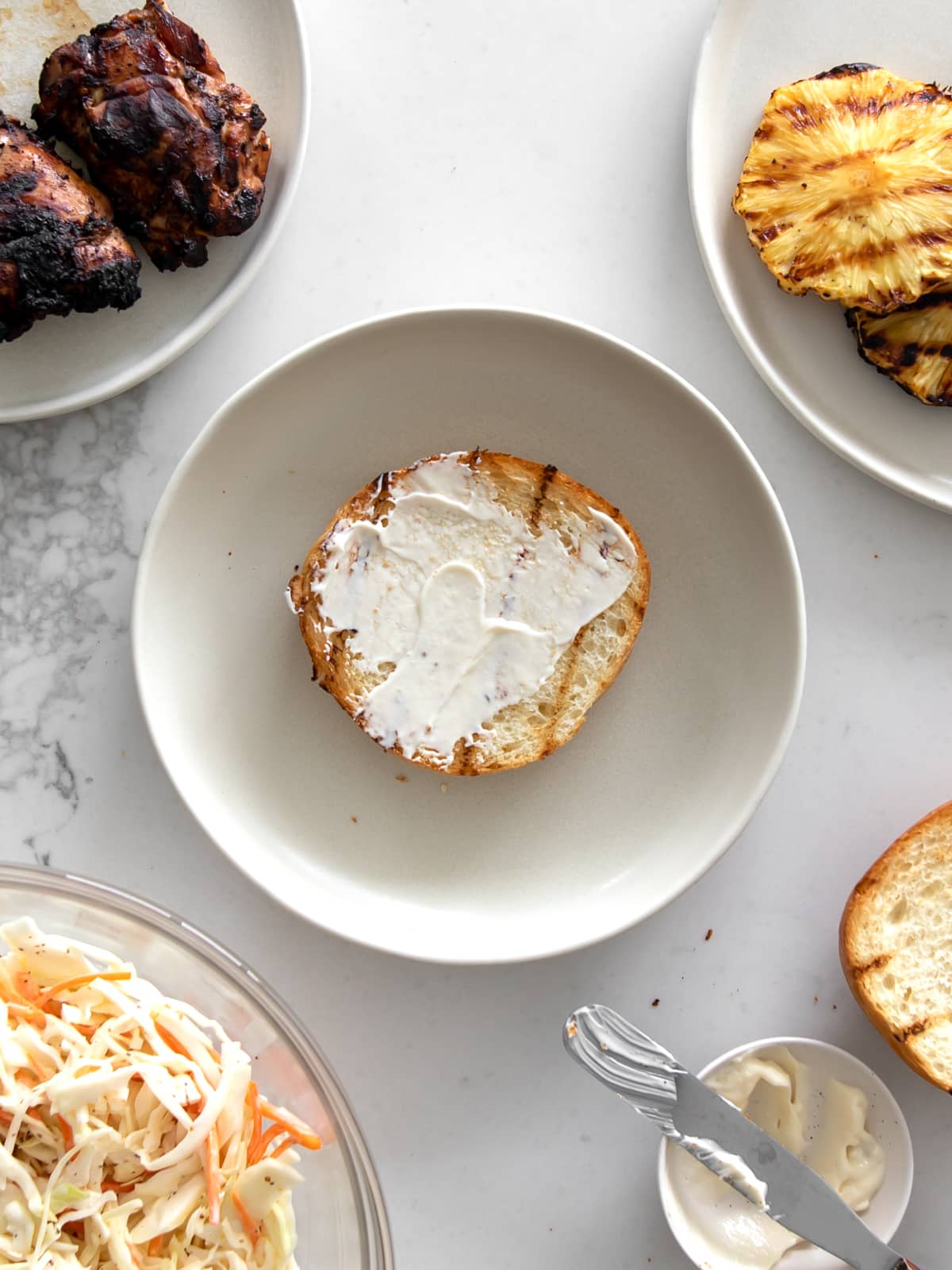 Toasted bun with mayonnaise on a plate, with plates of grilled jerk chicken, grilled pineapple, and coleslaw around it.