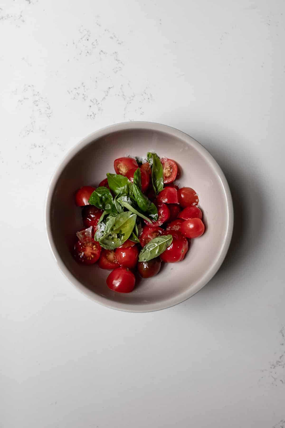 Fresh basil and grape tomatoes tossed in olive oil and salt in a grey bowl on a white marble background.