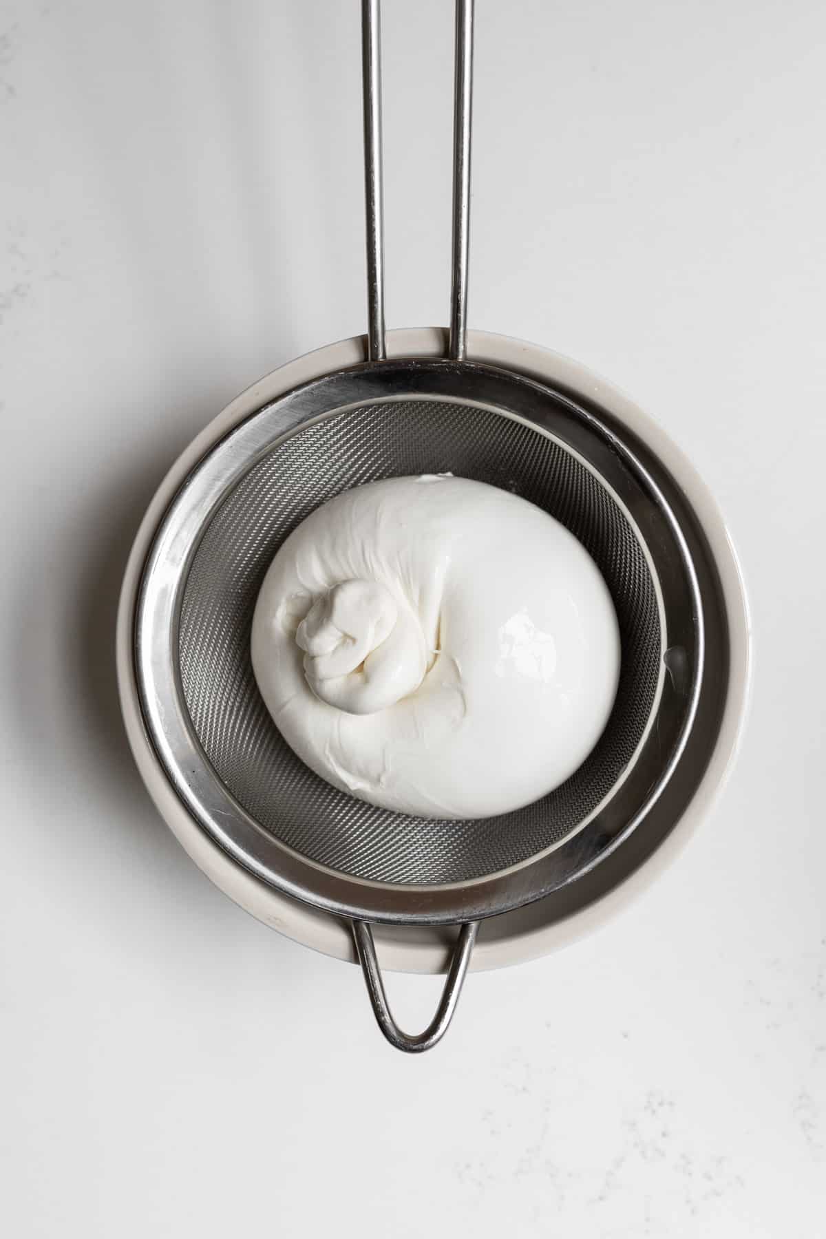 Ball of burrata cheese in a sieve placed over a beige bowl. 