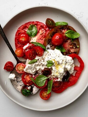 4 slices of heirloom tomatoes, grape tomatoes, small leaves, and spoonful of burrata cheese on a grey plate with antique serving spoon.