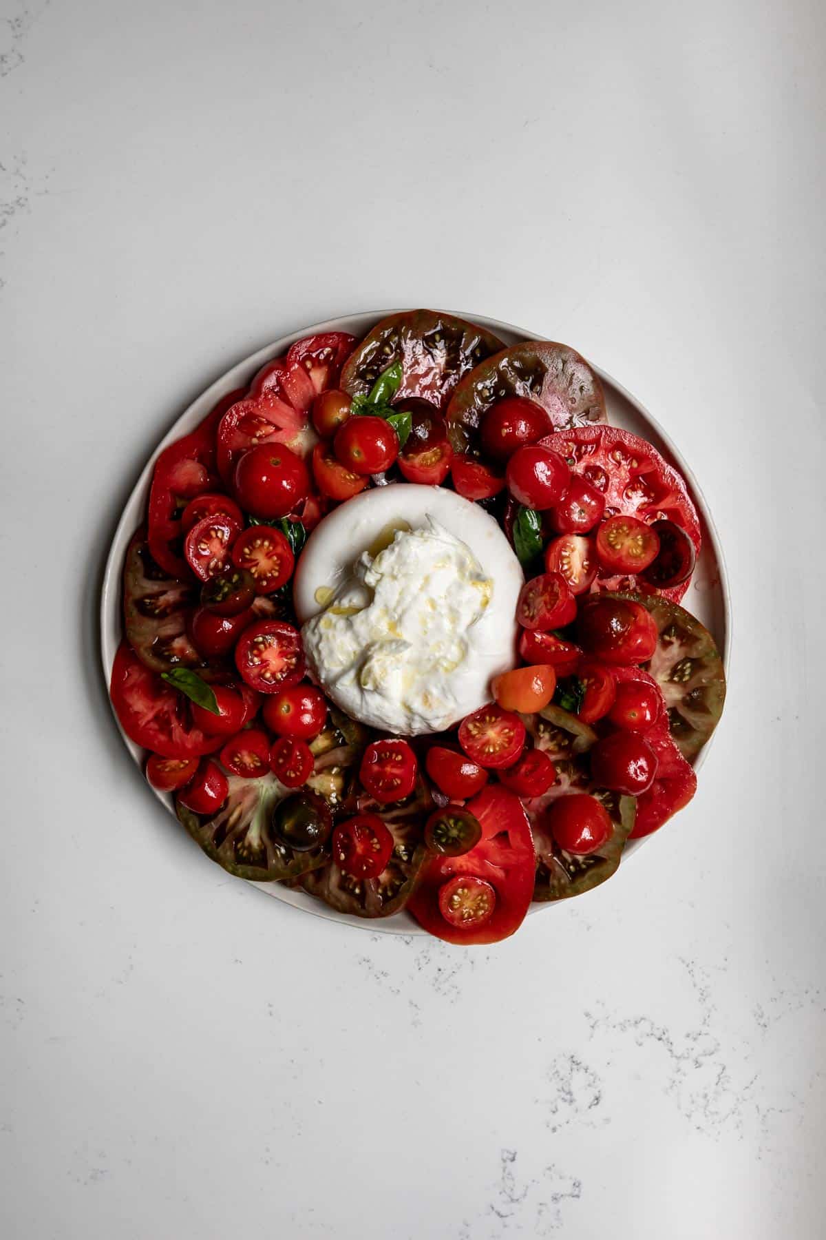 Ball of burrata cheese surrounded by sliced heirloom and grape tomatoes on a white plate with white marble background.