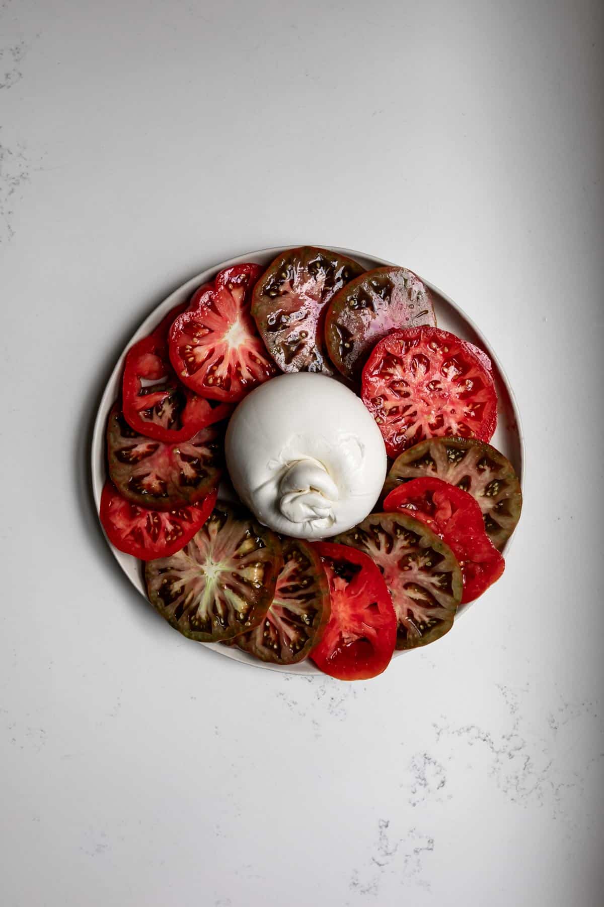 Ball of burrata cheese surrounded by sliced heirloom tomatoes on a white plate with while marble background.