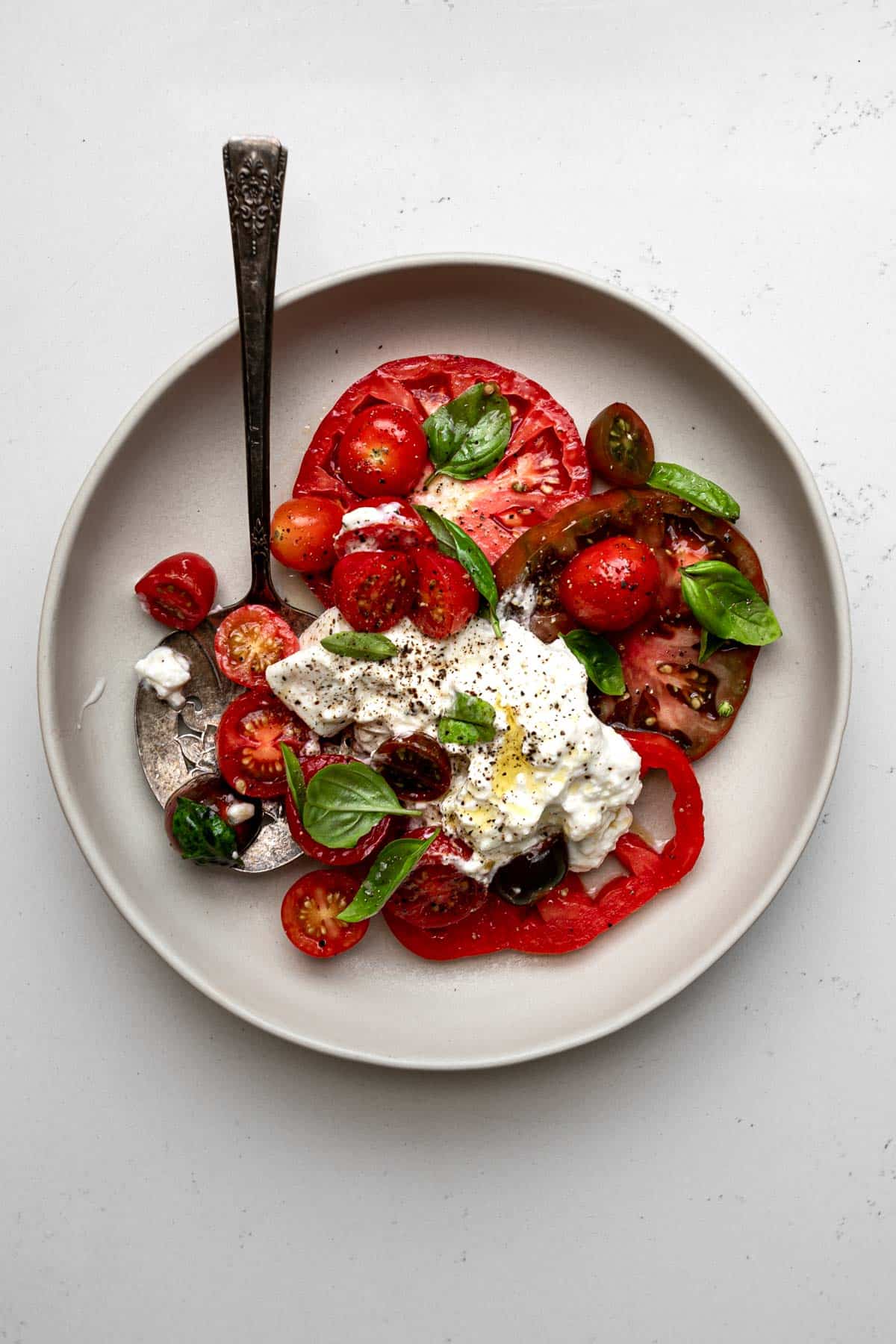 Plated burrata caprese with 4 slices of heirloom tomatoes, grape tomatoes, small leaves, and spoonful of burrata cheese on a grey plate with antique serving spoon.