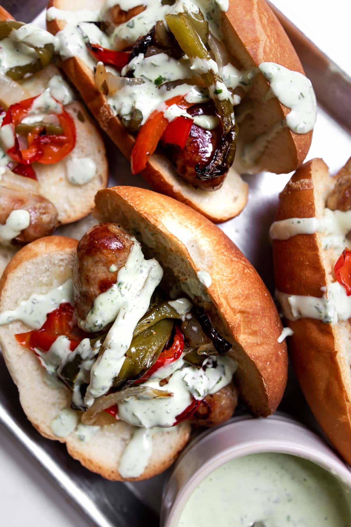 Close up of Italian sausage on a bun with roasted peppers, onions and basil mayonnaise sauce along with ramekin of basil mayo.